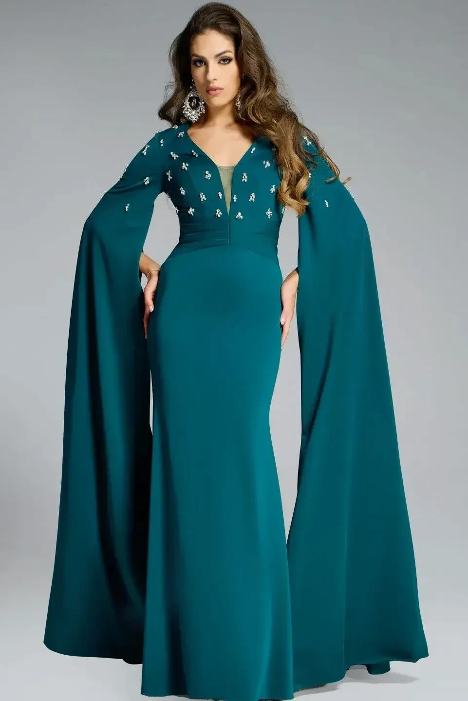A woman wearing a long, teal evening gown with long sleeves and embellishments on the bodice, standing in a graceful pose.