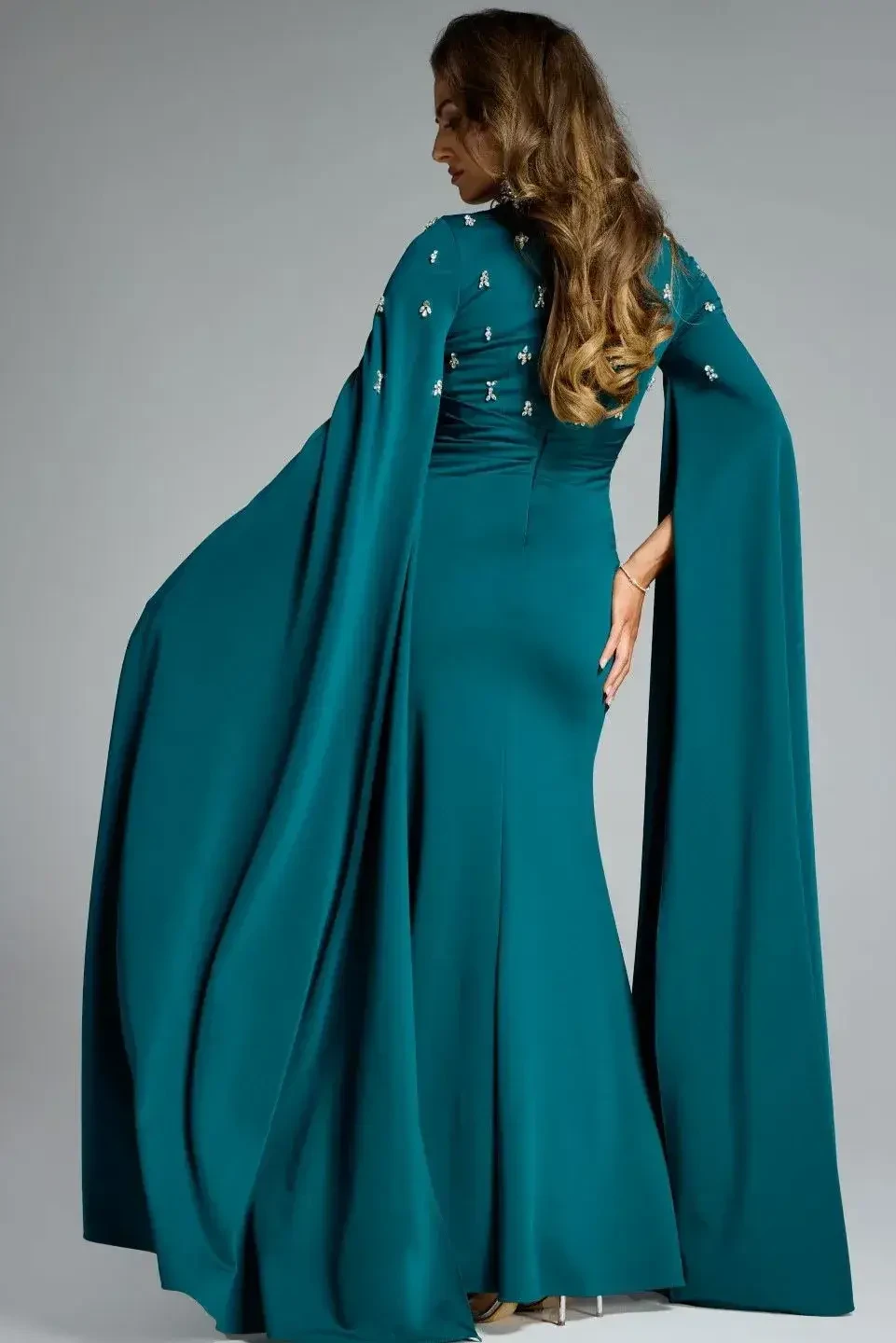 A woman wearing a long, teal evening gown with long sleeves and embellishments on the bodice, standing in a graceful pose.