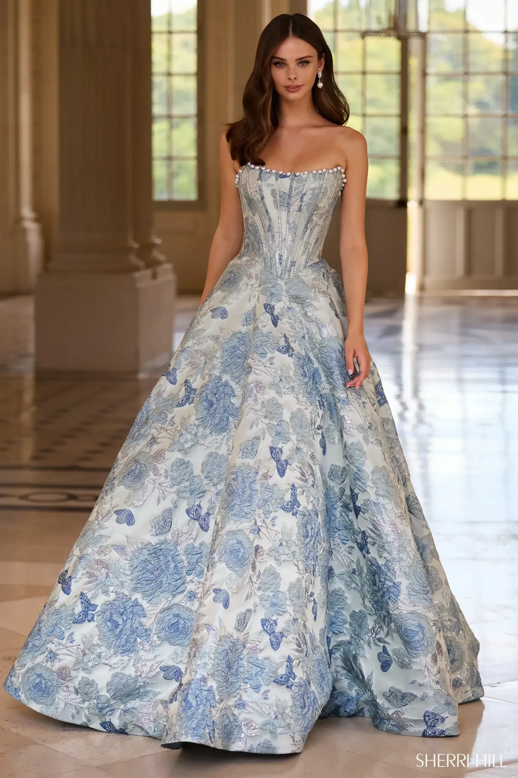 A model wearing a strapless, floral-patterned ball gown with shades of blue and silver, standing in an elegant indoor setting.