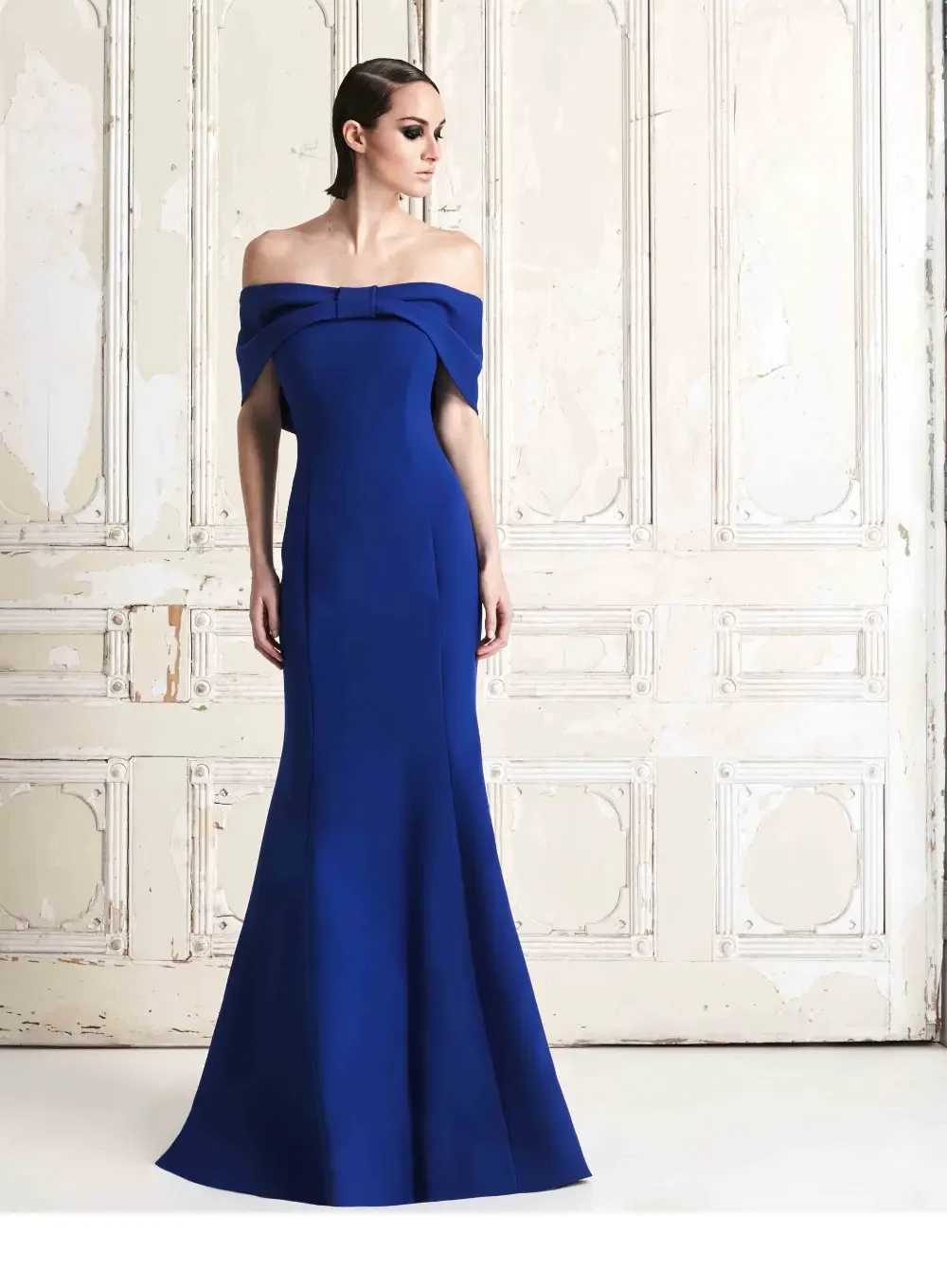 A model wearing a striking blue off-the-shoulder gown stands gracefully against a vintage backdrop with white, paneled walls.