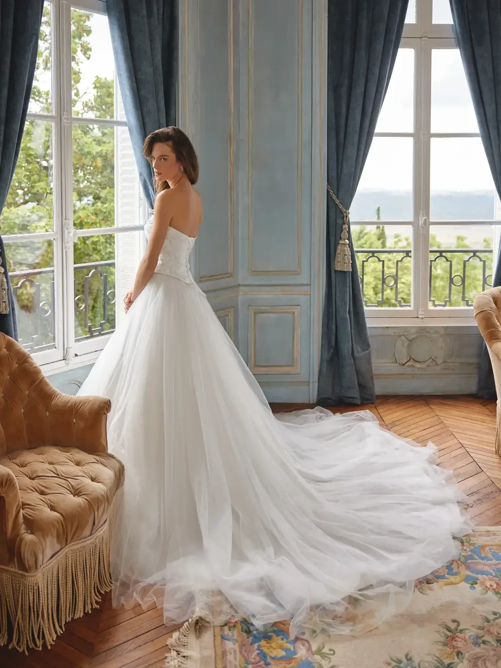 A woman in a strapless white wedding dress stands in an elegantly decorated room with blue curtains, a vintage lamp, and ornate furniture.