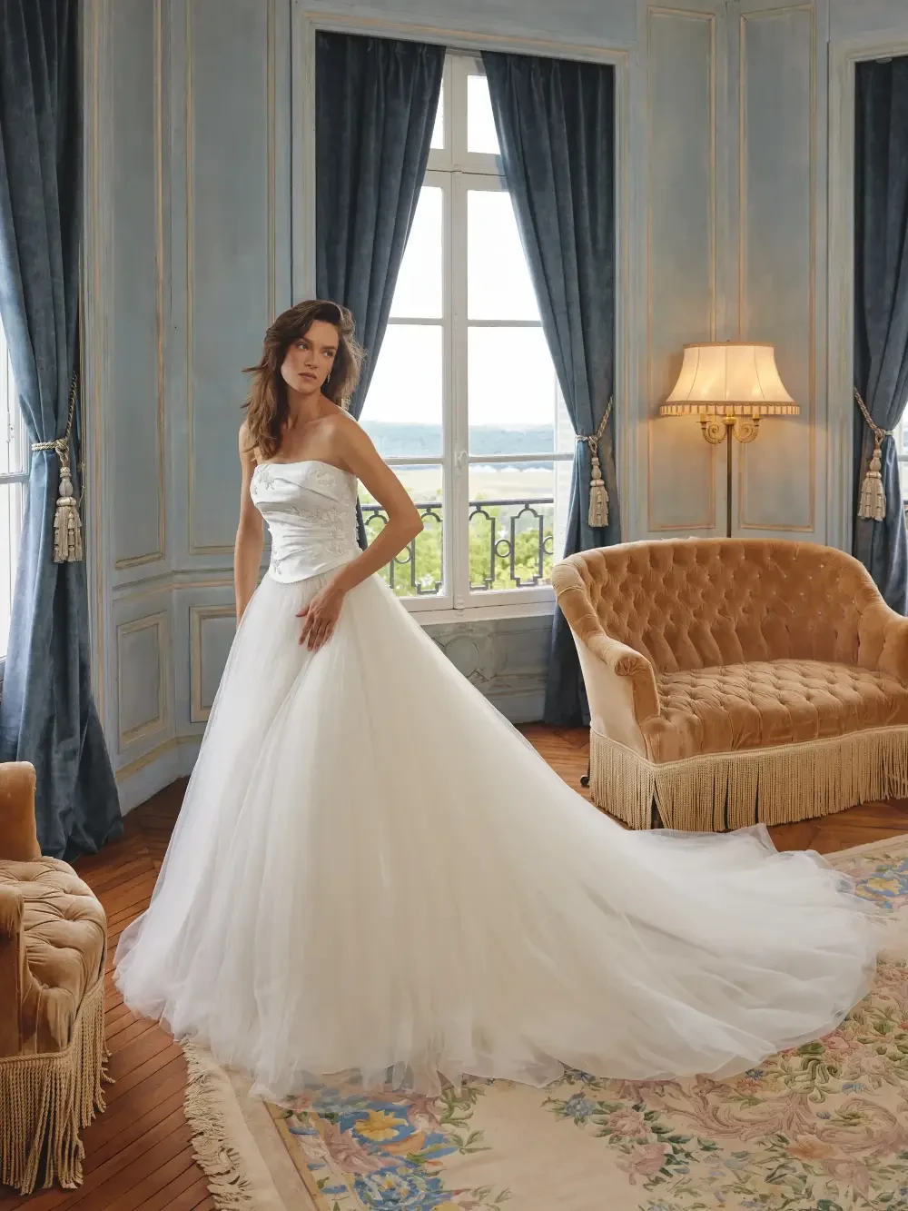 A woman in a strapless white wedding dress stands in a beautifully decorated room with large windows and elegant furnishings, including a beige sofa and curtains.