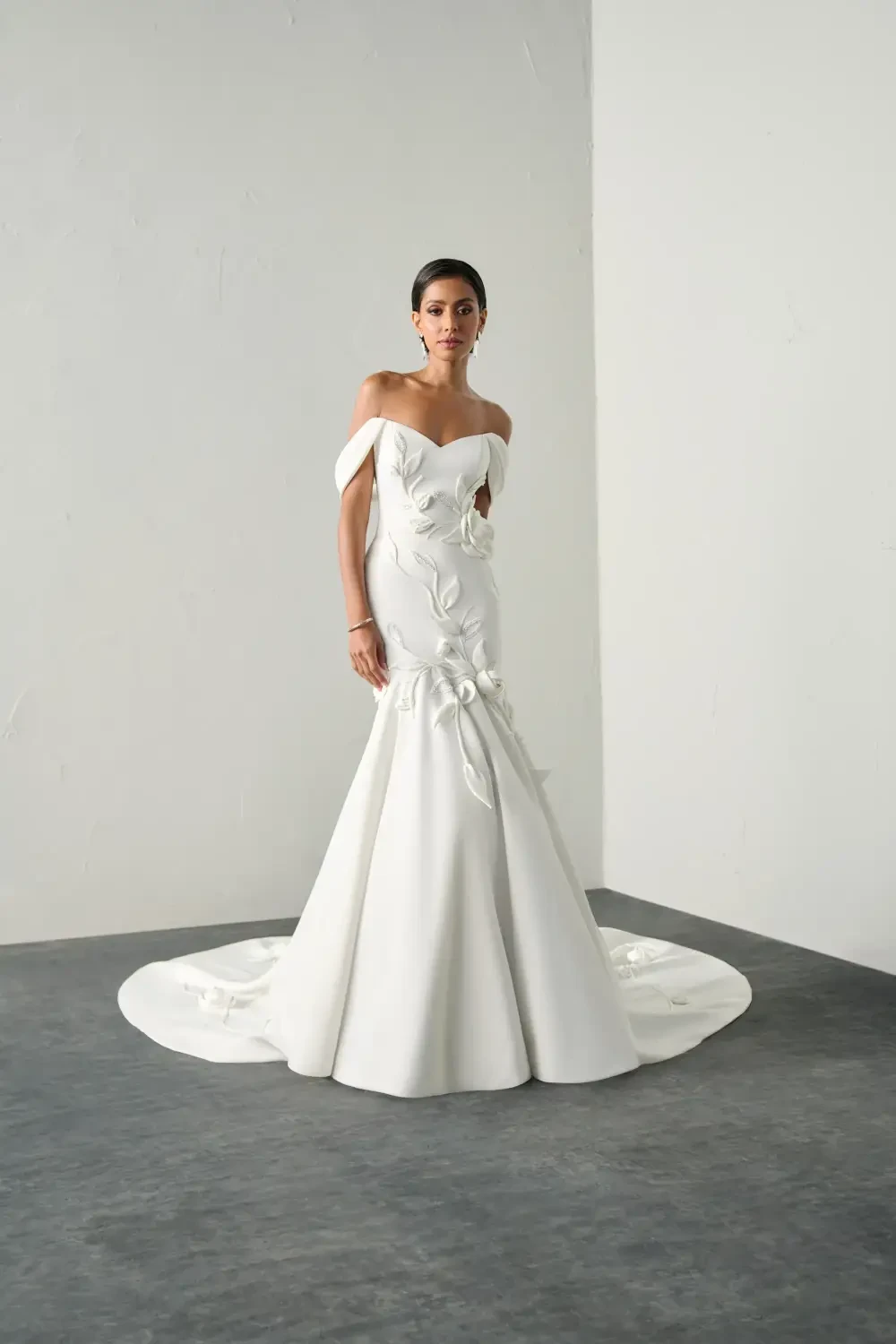 A bride wearing a stunning off-the-shoulder white wedding gown with floral details and a flowing train, standing in a minimalist setting.