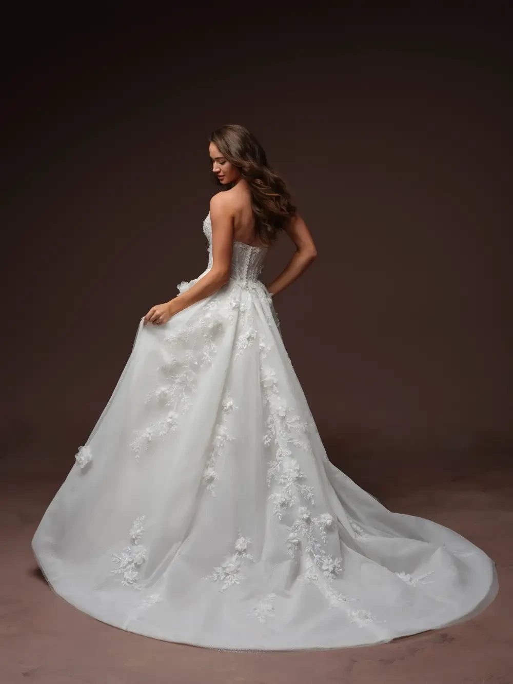 A woman wearing an elegant white wedding gown with floral details and a long veil, standing against a dark background.