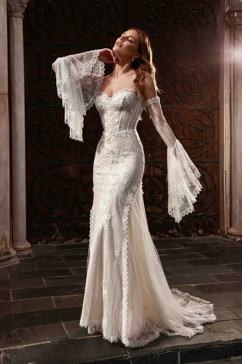 A woman posing in an elegant, intricately designed wedding dress with lace details and long, flowing sleeves, set against a decorative background.