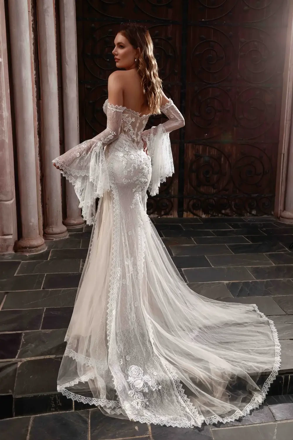 A woman posing in an elegant, intricately designed wedding dress with lace details and long, flowing sleeves, set against a decorative background.