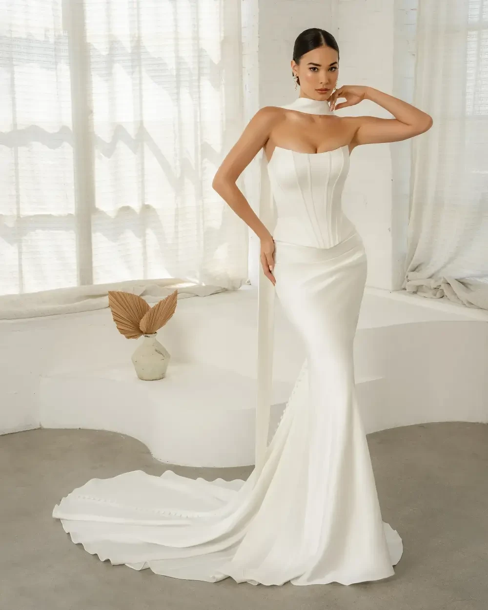A woman in a sleek, white wedding dress stands gracefully in front of a large window. The dress features a fitted bodice and a long train, emphasizing elegance and sophistication.